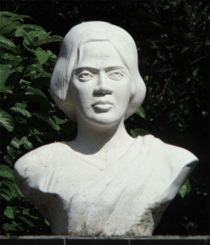 A white stone bust of Pritilata Waddedar with bobbed hair and a draped sari, set against dark green foliage.