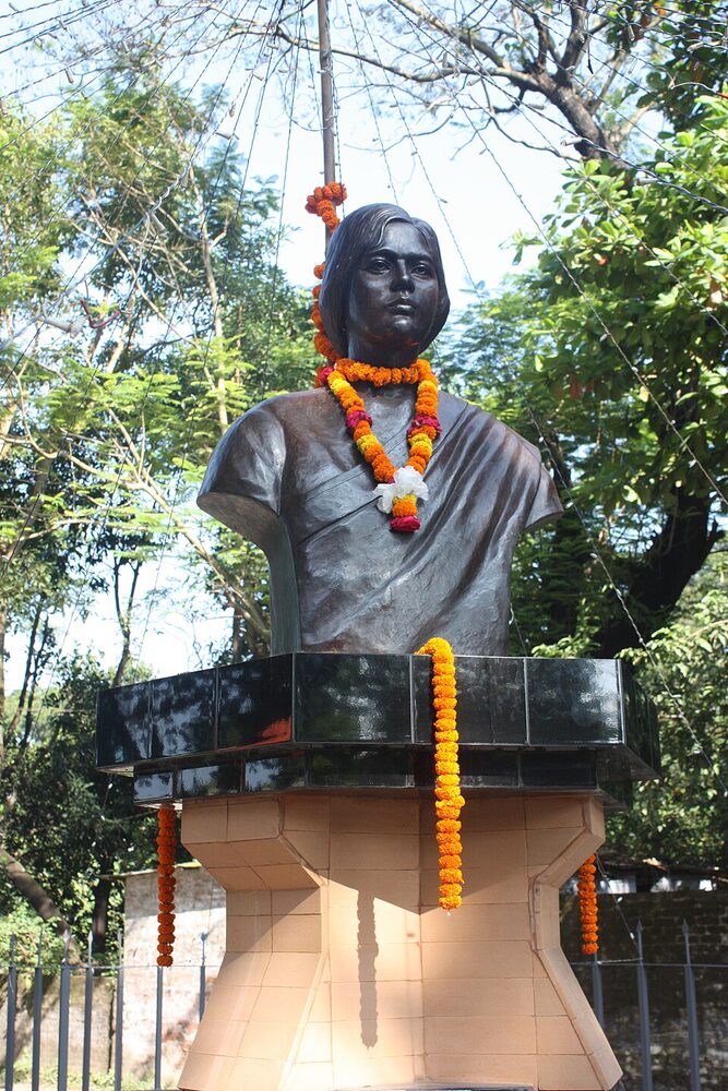 A dark bronze bust of Pritilata Waddedar on a stepped plinth, hung with marigold garlands, standing outdoors among trees.