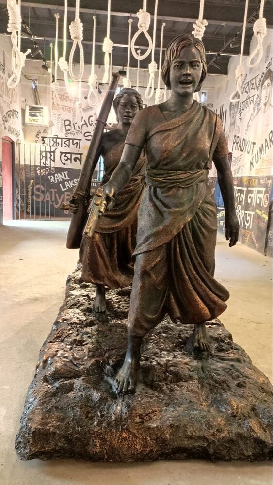 A bronze-toned sculptural group of two women in saris striding forward, displayed in a museum gallery whose walls carry the painted names of revolutionaries.