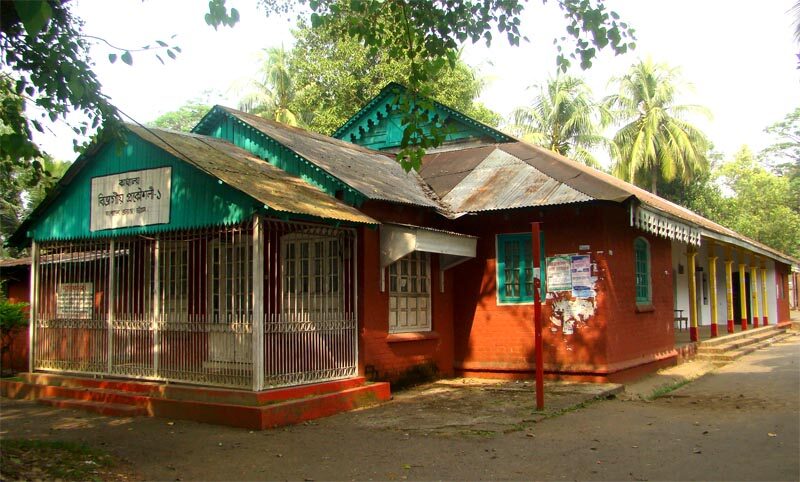 A low colonial-period building with a green corrugated roof and red walls, among palms and trees.