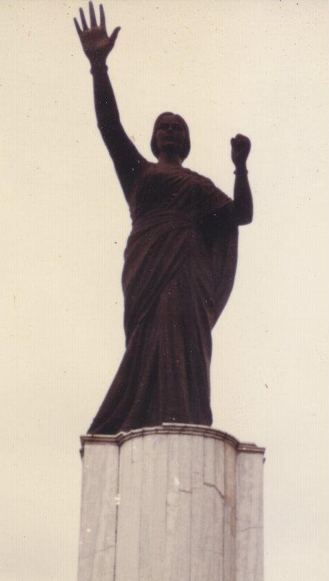 A dark full-length statue of a woman in a sari with her right arm raised and left hand clenched, on a tall fluted pedestal against a pale sky.